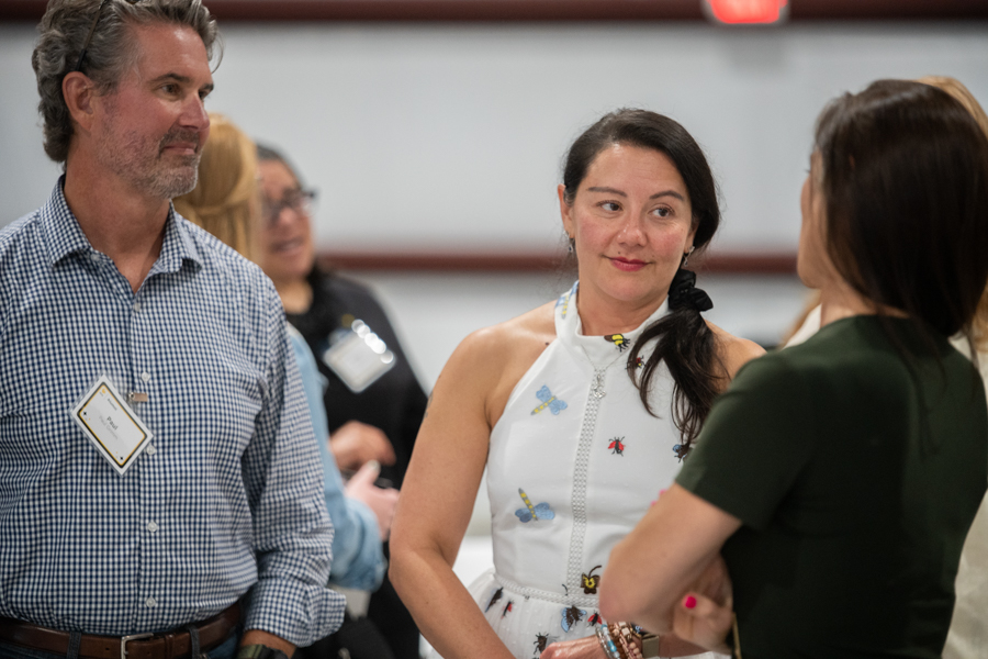 Woman in a white sleeveless dress talks with a colleague in green while a man with a name badge listens nearby at a social/professional event.