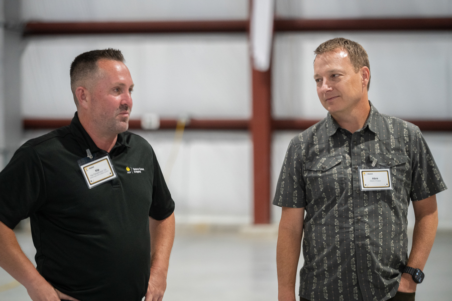 Two men with name badges talk in a large indoor warehouse setting, facing each other in conversation.