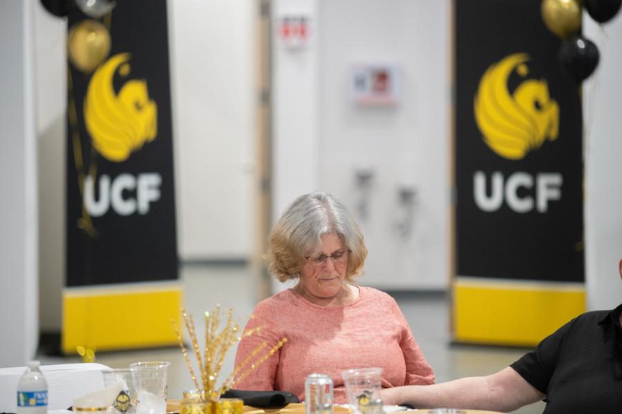 Older woman with gray hair wearing a pink sweater sits at a table during a UCF event, with black-and-yellow UCF banners in the background.