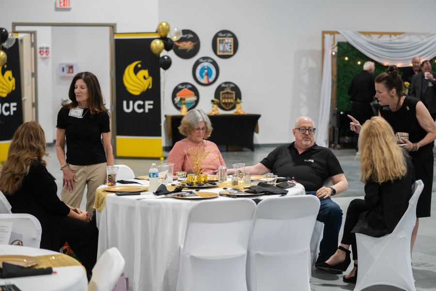 Group of adults seated around a round banquet table with black and gold place settings at an indoor event; UCF banners and decorations visible in the background.