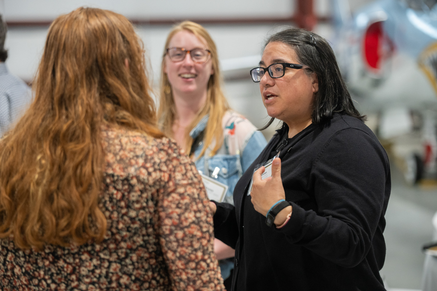 Three women conversing in a workshop with airplane parts visible; the woman in a black cardigan speaks while the others listen.