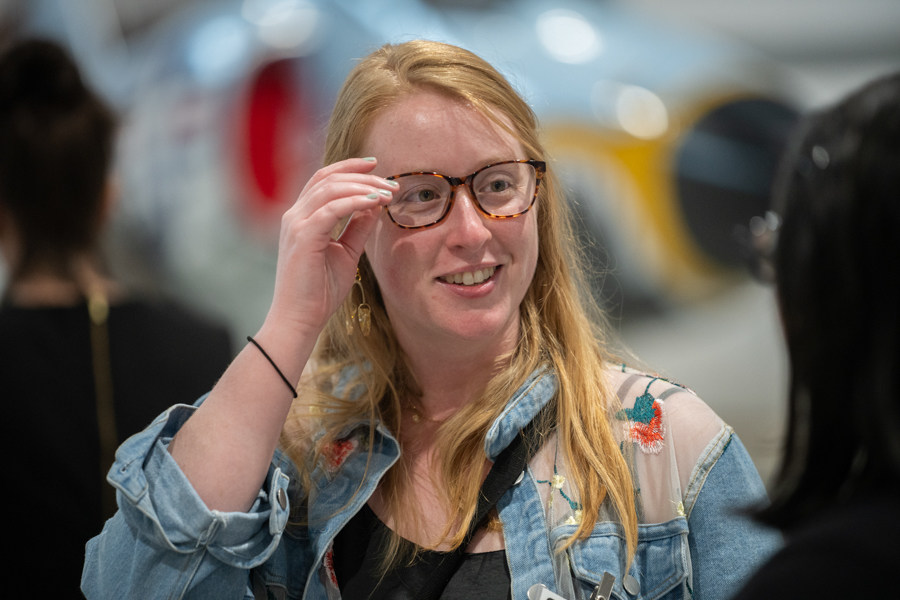 Woman with blonde hair and tortoiseshell glasses smiles while adjusting her glasses in a denim jacket with floral embroidery at an indoor event.
