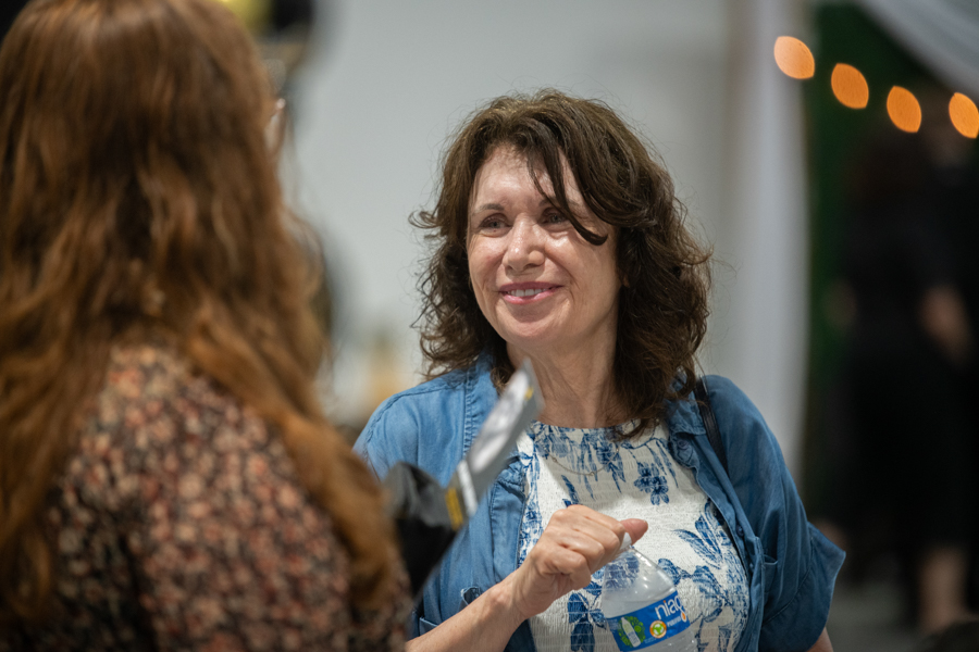 Smiling woman in a blue shirt holds a water bottle while talking to another person at an indoor event.
