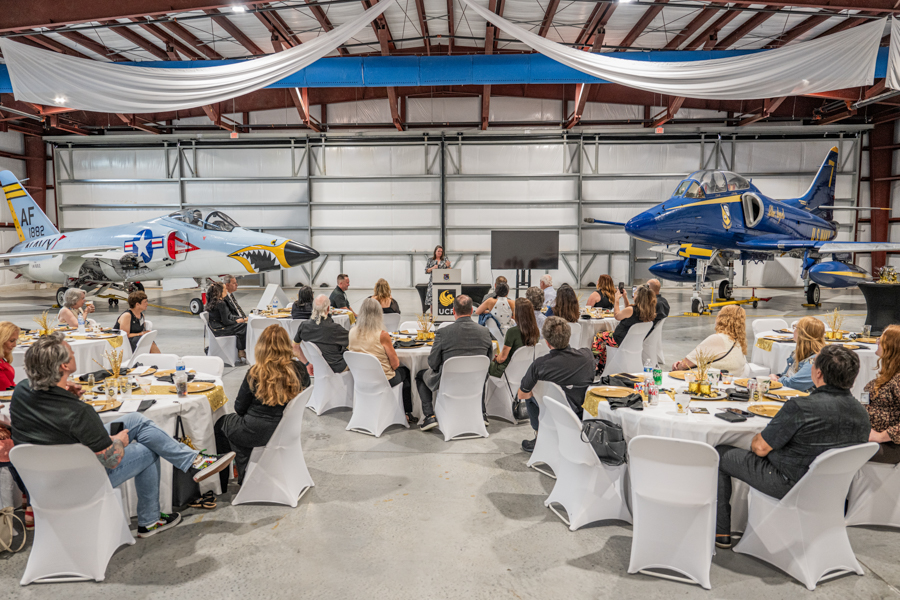 Formal dinner in an airplane hangar: guests at round tables while a speaker presents at a podium, with aircraft on display.