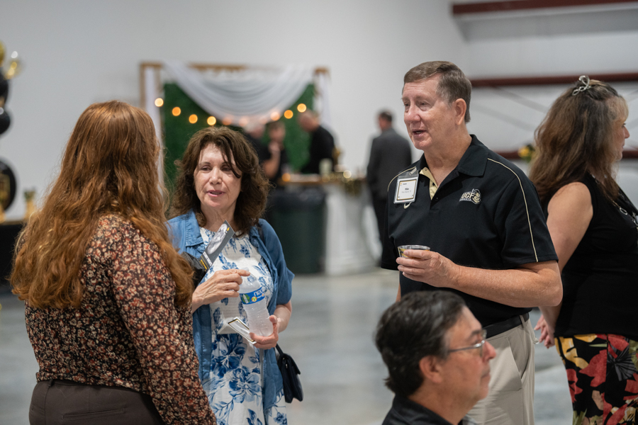 Three adults chat at a reception inside a spacious hall; a woman in a blue floral dress and denim jacket holds a water bottle while a man in a black polo speaks with a drink in hand.