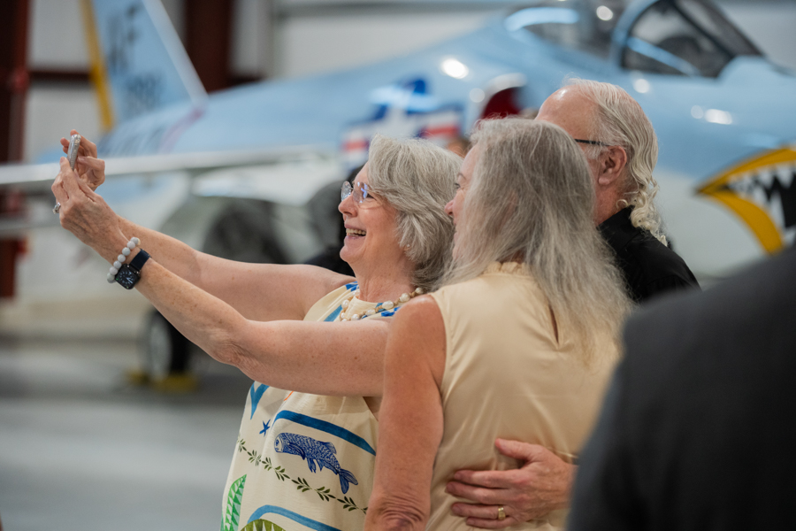 Smiling elderly woman holds up a phone to take a selfie with two friends in a hangar, a light-blue aircraft with shark-face art in the background.