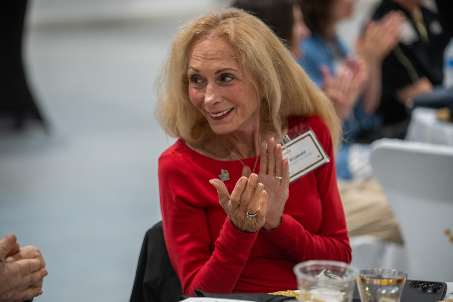 Smiling blonde woman in a red top clapping at a table during a formal event, wearing a name badge.