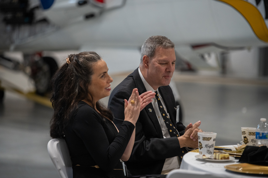 Woman and man in formal attire clap at a table with coffee cups inside an airplane hangar with a parked plane in the background, watching an event in progress.
