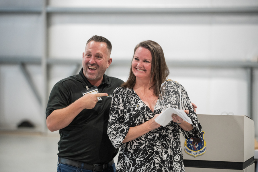 Two smiling speakers stand at a podium; man points to the papered woman as they pose for a photo at an event.
