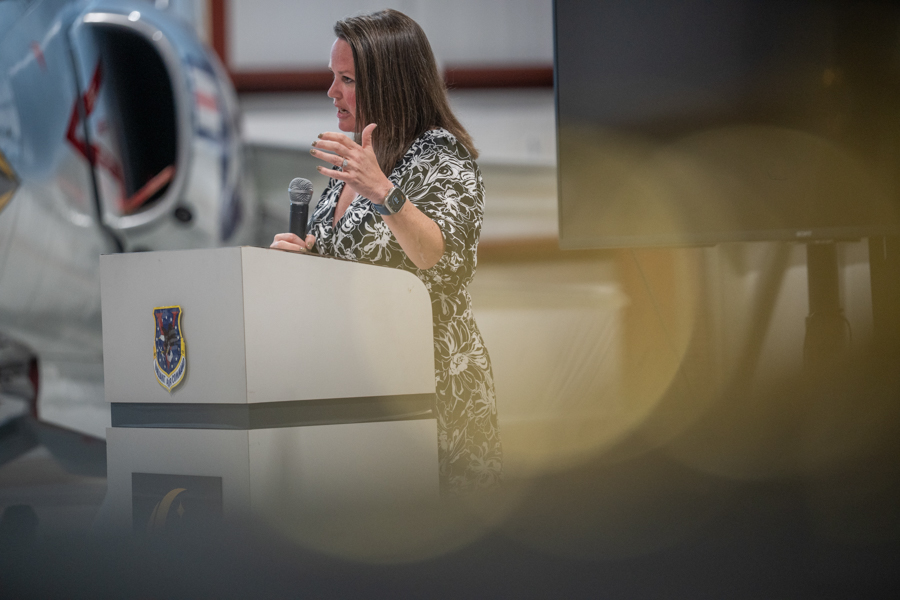 Woman speaking at a podium with a microphone, in front of a small aircraft in a hangar, emblem on the podium visible