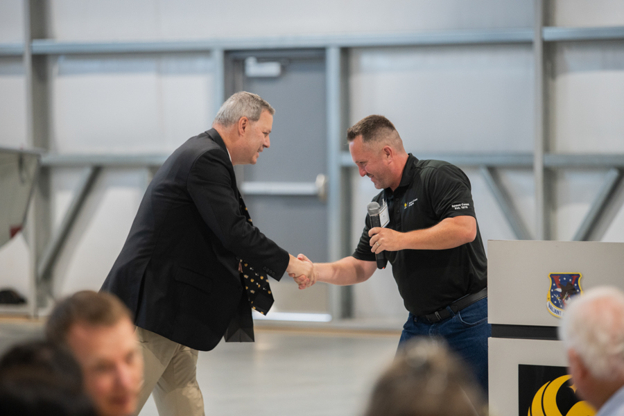 Two men shake hands on a stage during a formal event; one holds a microphone as they greet each other in front of a podium.