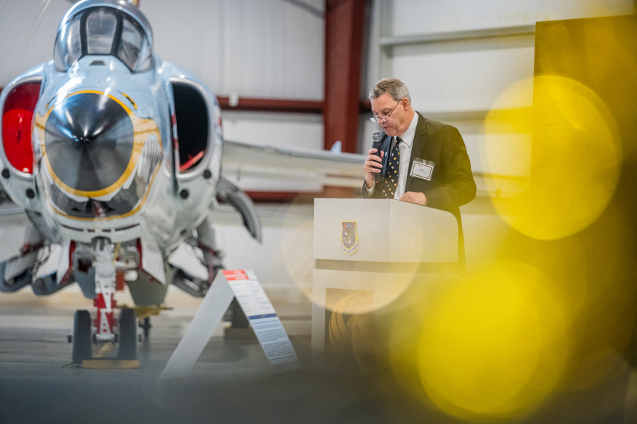 Man in a suit giving a speech at a podium beside a jet aircraft in a hangar, with yellow foreground blur in the foreground.