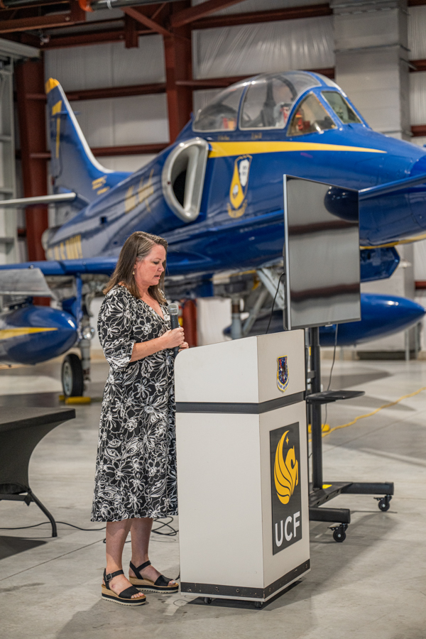 Woman in a patterned dress speaks into a microphone at a lectern with a UCF logo, in an airplane hangar with a blue jet in the background.