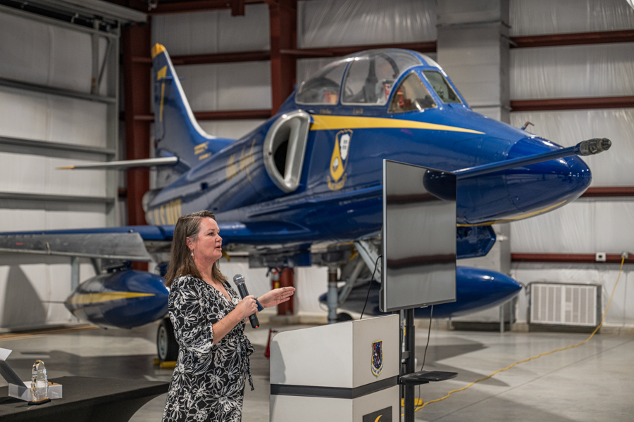 Woman speaking at a podium with microphone in front of a blue jet inside a hangar