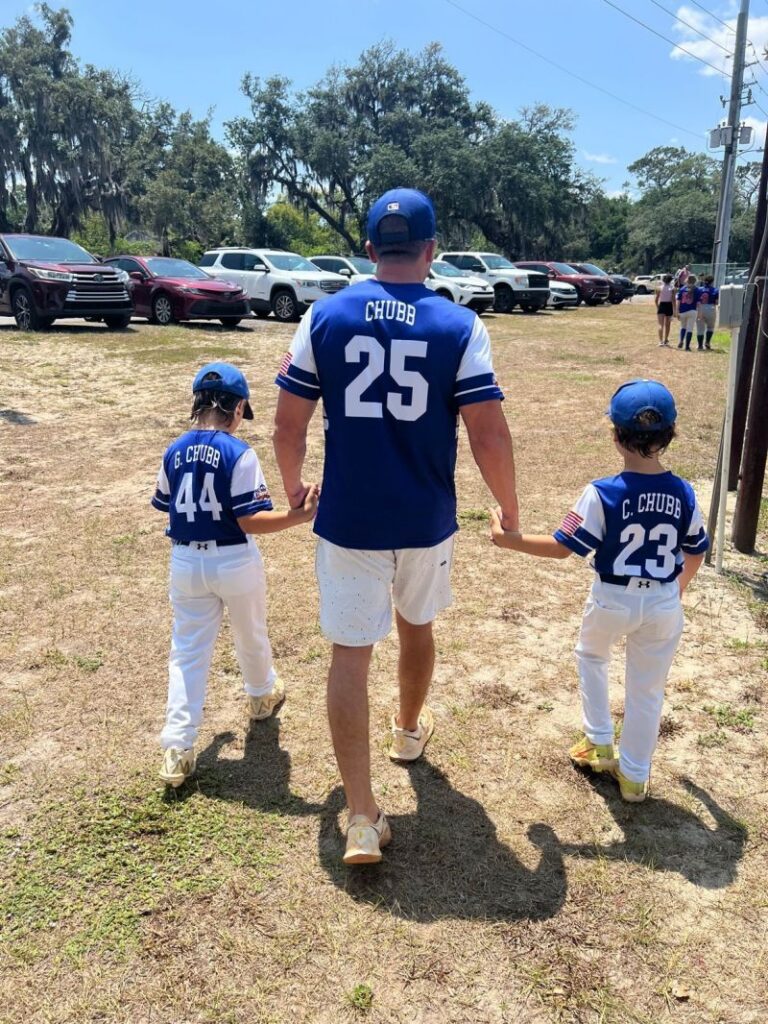 Man in blue baseball jersey number 25 walks hand-in-hand with two children in matching blue and white uniforms on a sunny day, with cars in a parking lot in the background.