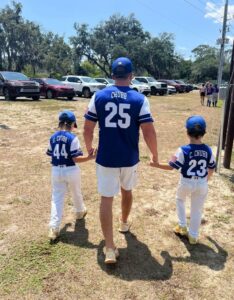 Man in blue baseball jersey number 25 walks hand-in-hand with two children in matching blue and white uniforms on a sunny day, with cars in a parking lot in the background.
