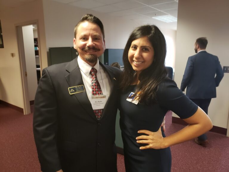 Two professionals pose together in a hallway, both wearing name badges at a conference or workplace event.