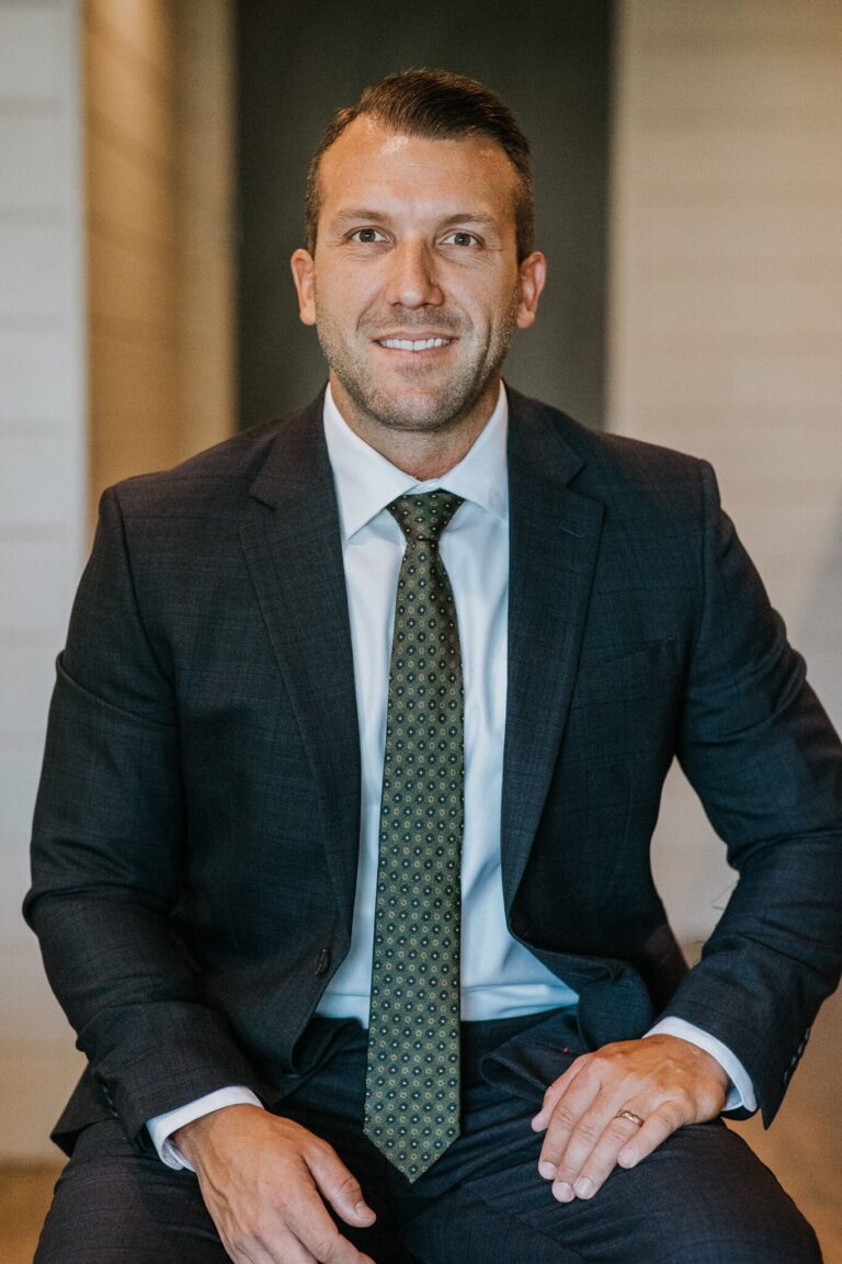 Head-and-shoulders portrait of a smiling man in a dark suit and green patterned tie, seated indoors with a neutral background.