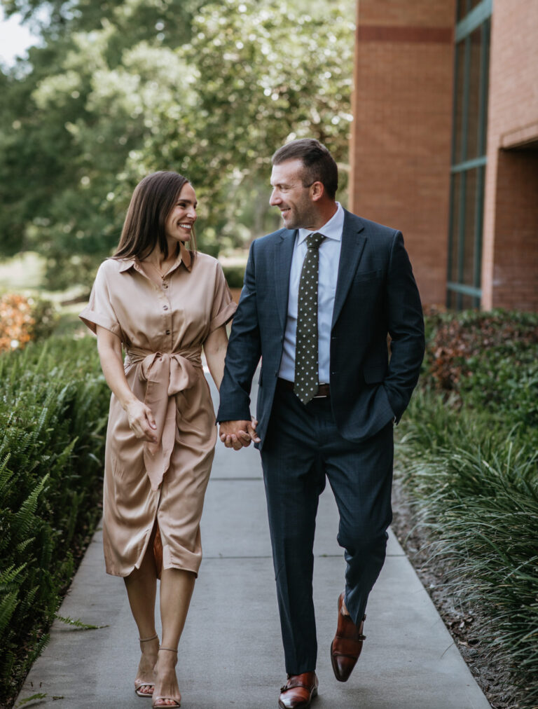 A couple in business attire walks hand in hand along a sidewalk, smiling at each other outdoors near a brick building.