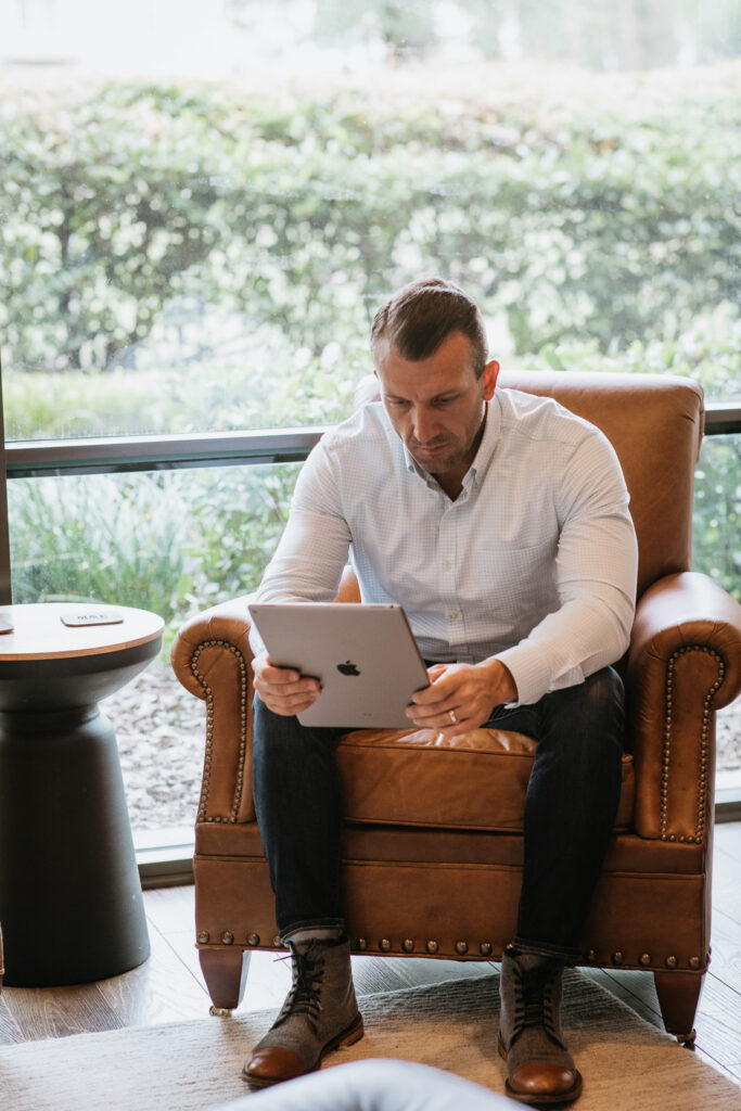 Man in a light shirt sits in a brown leather armchair, looking down at a tablet he's holding.