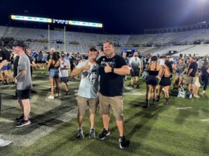 - UCF Alumni Two men on a football field at night pose together for a photo, smiling and giving thumbs up amid a crowded stadium backdrop.