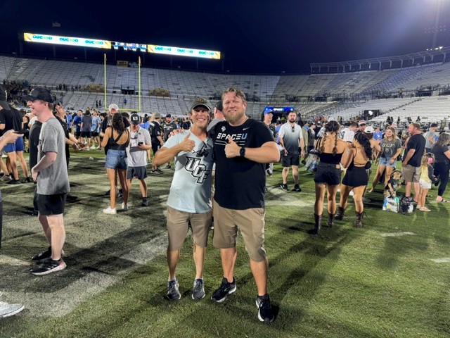 Two men on a football field at night pose together for a photo, smiling and giving thumbs up amid a crowded stadium backdrop.