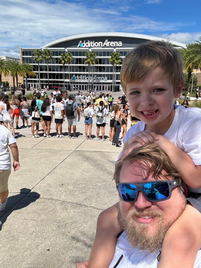 Man and child taking a selfie in the foreground outside Addition Arena, with a crowd gathered and a glass-front arena building behind them on a sunny day.