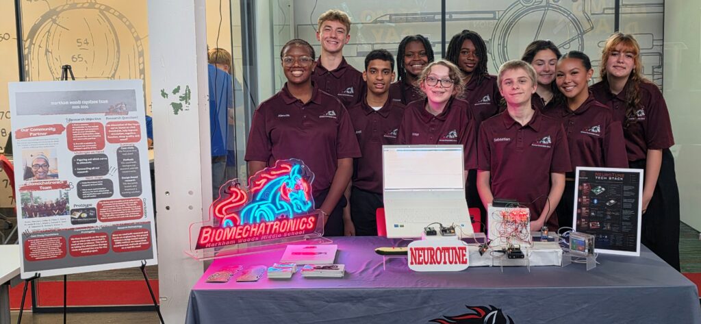 Group of students in maroon shirts posing at a Biomechatronics booth with posters and laptops behind them.