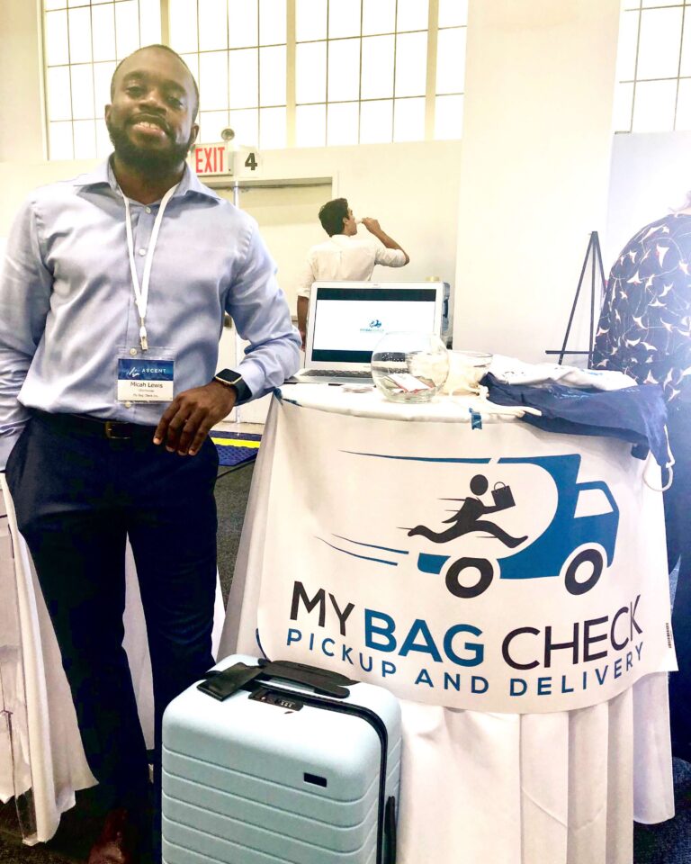 Smiling man in a blue shirt with a name badge at a booth, next to a banner reading 'My Bag Check' with a blue suitcase on the table nearby.
