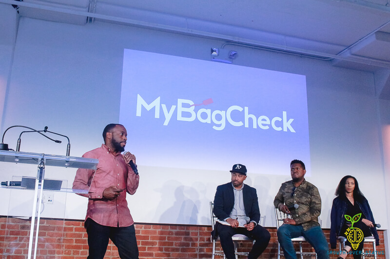 Panel discussion on a stage with four people; a projection shows the 'MyBagCheck' logo behind them.