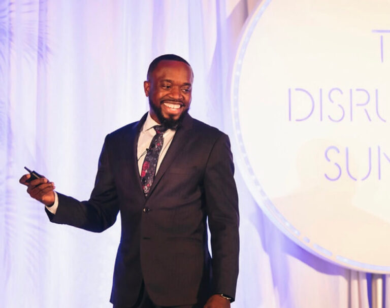Smiling man in a dark suit presenting on stage, holding a remote in his right hand with a circular backdrop that reads 'DISR…' behind him.