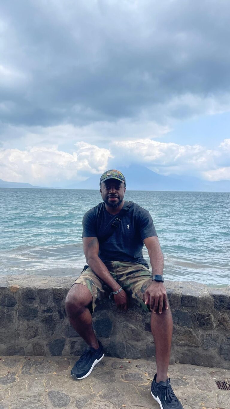 Man sitting on a stone seawall by the water, wearing a cap, dark T-shirt, camouflage shorts, and sneakers; cloudy sky overhead.
