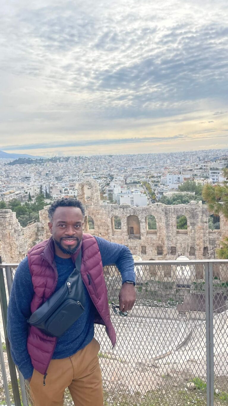 Smiling man in a burgundy vest and navy shirt stands at a railing in front of ancient stone arches with a cityscape in the background.