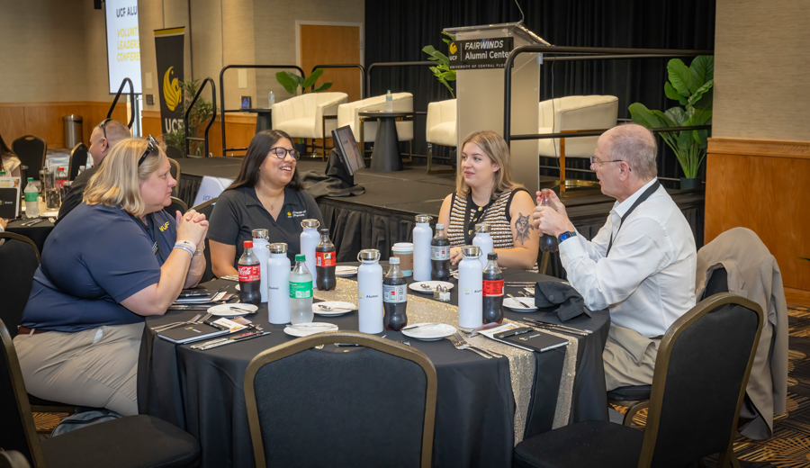 Four people sit around a round table chatting at a conference, with bottled drinks and plates on the table; stage with seating in background.