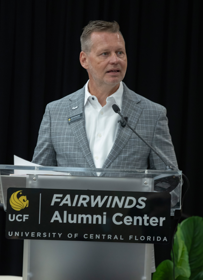 Man in a gray checkered blazer speaks at a podium branded with 'Fairwinds Alumni Center' and 'University of Central Florida' signage.