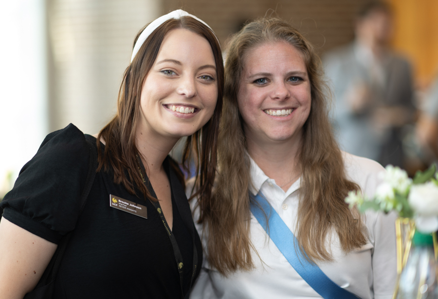 Two smiling women at a social event posing for a photo, one with a name badge on black outfit.