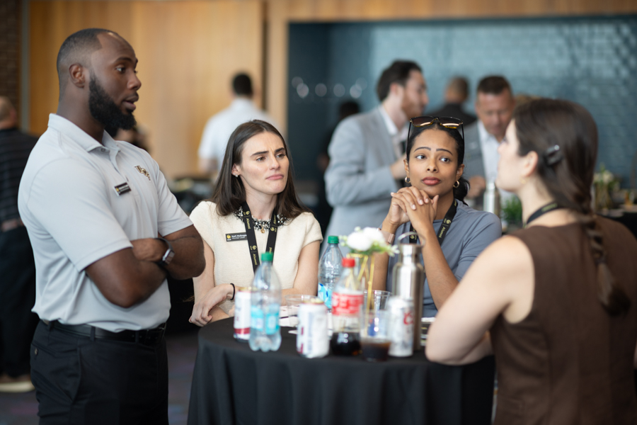 Man in light polo standing with arms crossed talking to three women seated at a round high-top table during a networking event, with drinks on the table