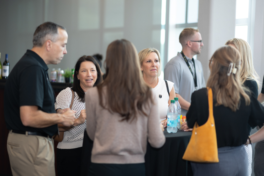 Group of adults talking around a high-top table at a networking event, with drinks visible on the table and in the background room.