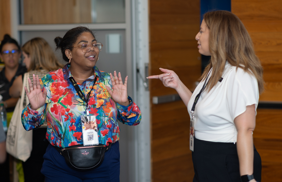 Two women converse in a hallway: one in a bright floral shirt with a waist bag, the other in a white blouse gesturing with her finger pointing.