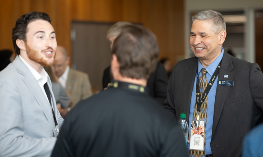 Two men in business attire chat at a conference, one facing camera in a light gray suit and the other with his back to us, smiling attendees nearby.