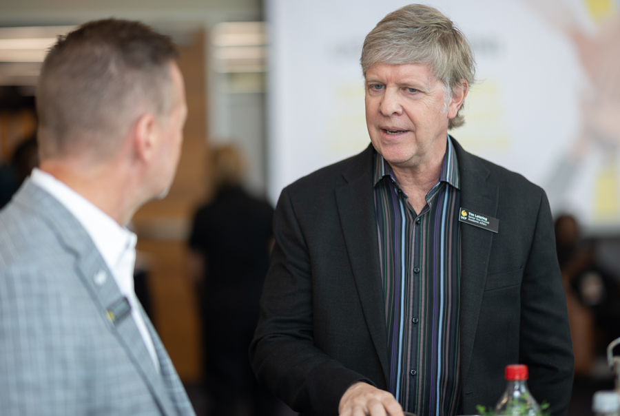 Two men in a business setting talk at a networking event; one wears a black blazer and name tag, the other in a gray suit.