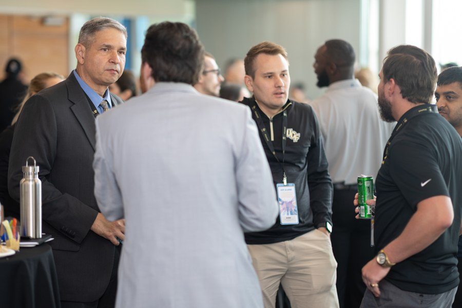 Group of professionals chatting at a networking event, wearing badges, with a high-top table and drinks nearby.