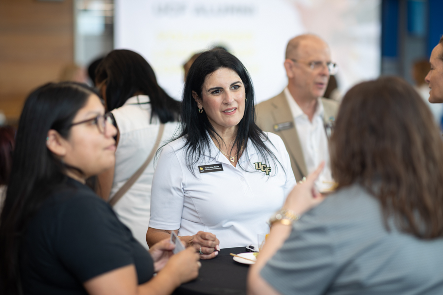Professional woman wearing a light blue polo with UC branding speaks with a group at a networking event around a high-top table.