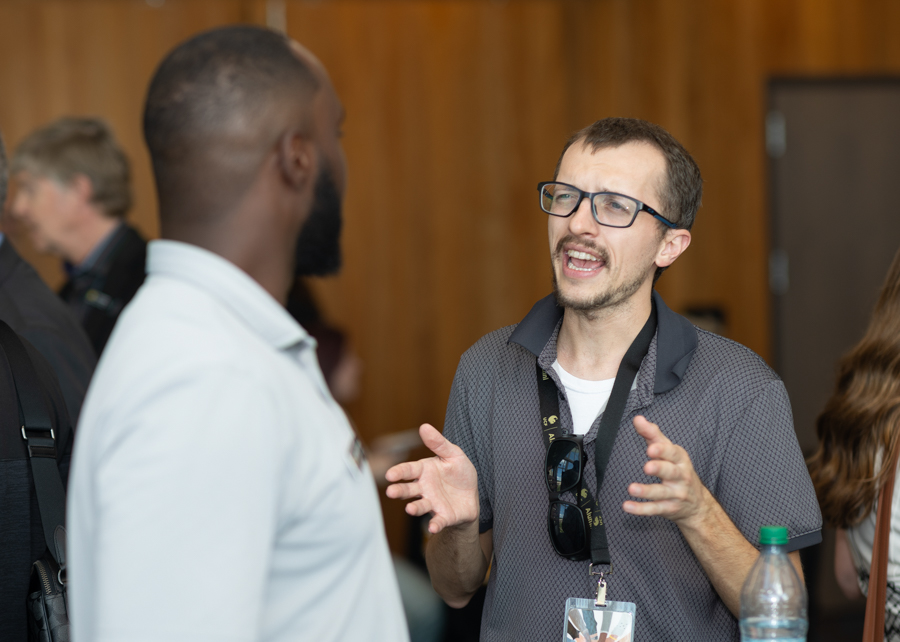 Man wearing glasses speaks with animated hand gestures to another man at an indoor event, wearing a lanyard and sunglasses hanging from his shirt.