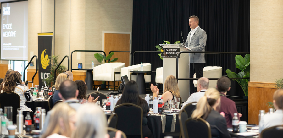 Man in a gray blazer giving a presentation at a podium labeled Fairwinds Alumni Center, in front of an audience seated at round tables in a conference room.