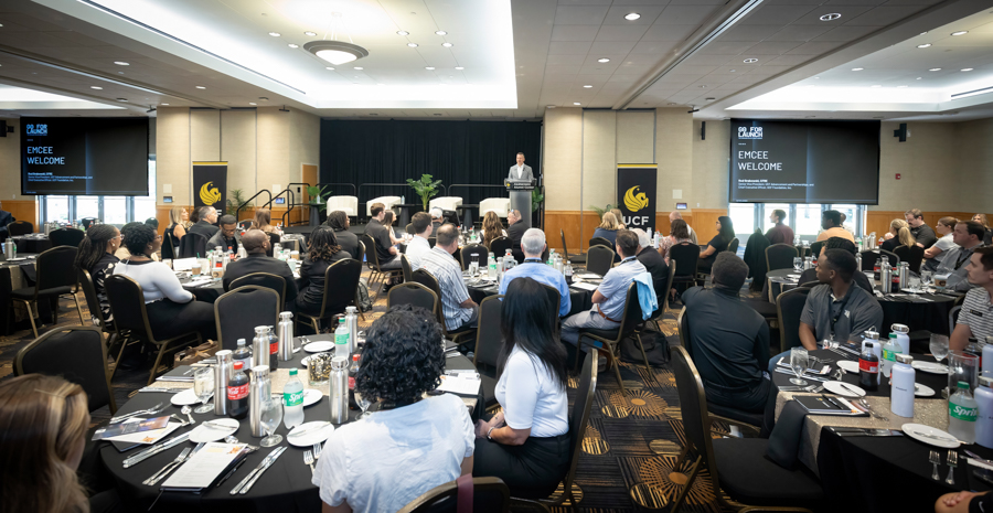 Speaker at a podium addressing a large audience in a conference hall with round tables and banquet-style setup