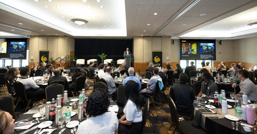 Speaker at a podium addressing attendees in a large conference room with round tables and screens on either side.