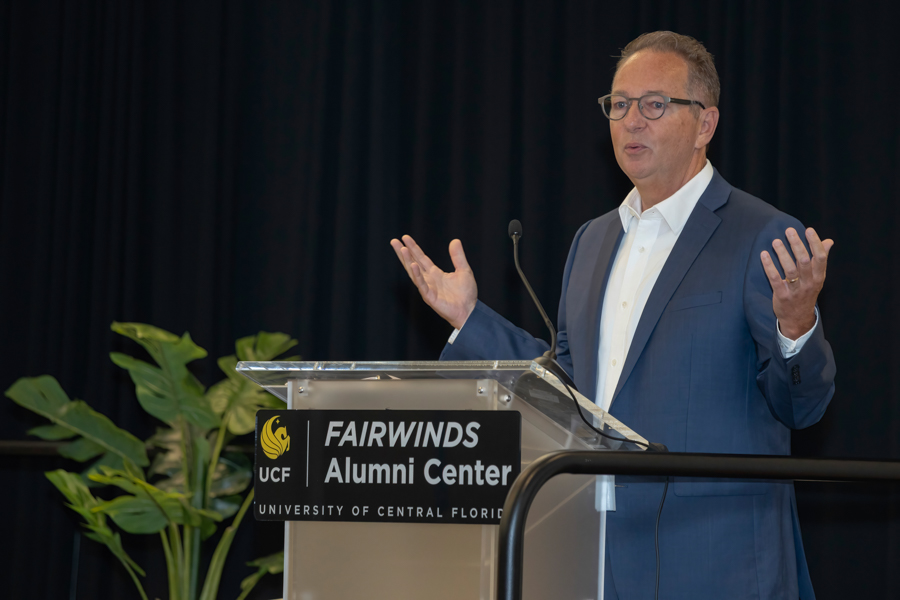 Man in a blue suit speaks at a podium labeled Fairwinds Alumni Center, University of Central Florida, with a microphone and a plant in background.