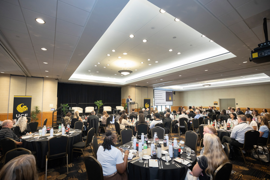Conference hall with a speaker at a podium addressing a round-table audience; banners with UCF logos on stage left and right.