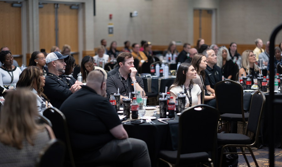 Audience seated at round tables in a conference hall, listening to a presentation at a professional event.